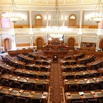 Vista ampla e vazia de uma câmara legislativa ornamentada, com cadeiras e lustres.