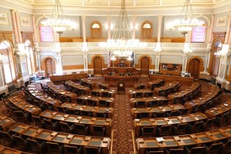 Vista ampla e vazia de uma câmara legislativa ornamentada, com cadeiras e lustres.