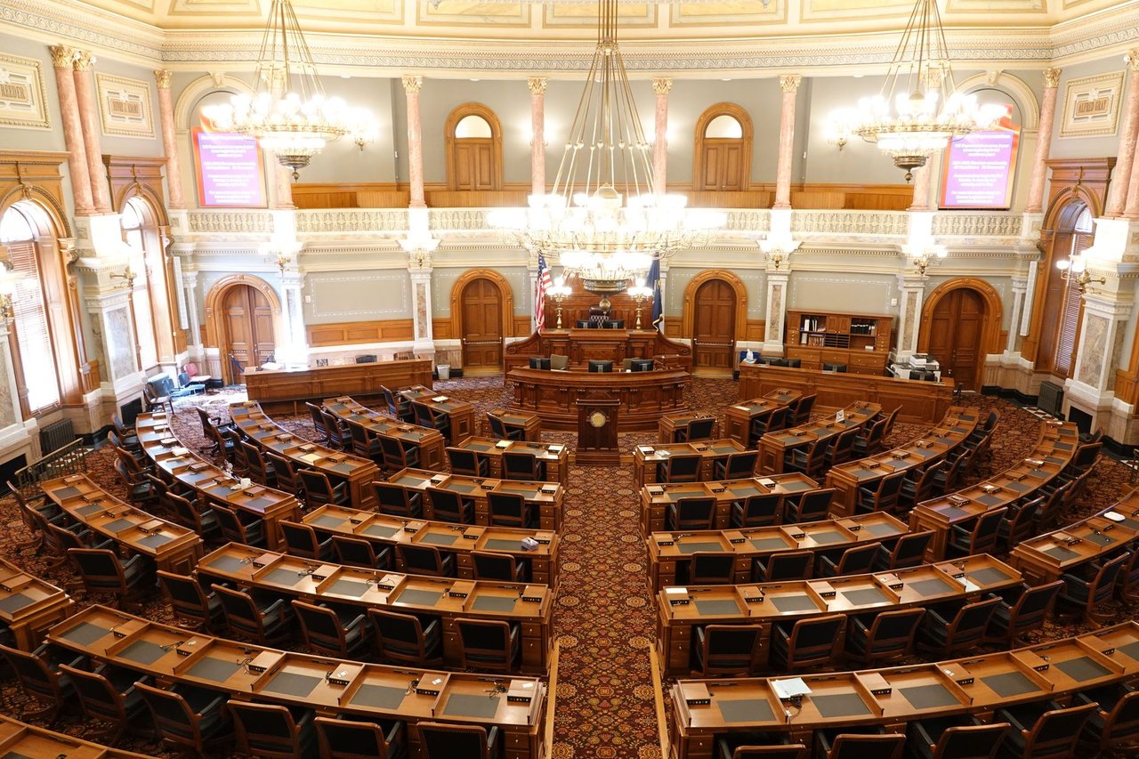 Vista ampla e vazia de uma câmara legislativa ornamentada, com cadeiras e lustres.