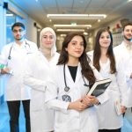 Grupo diversificado de estudantes de Medicina usando jalecos e conversando em um corredor universitário.