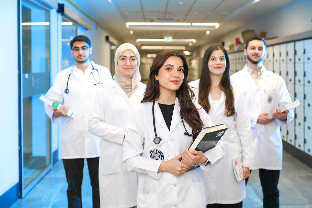 Grupo diversificado de estudantes de Medicina usando jalecos e conversando em um corredor universitário.